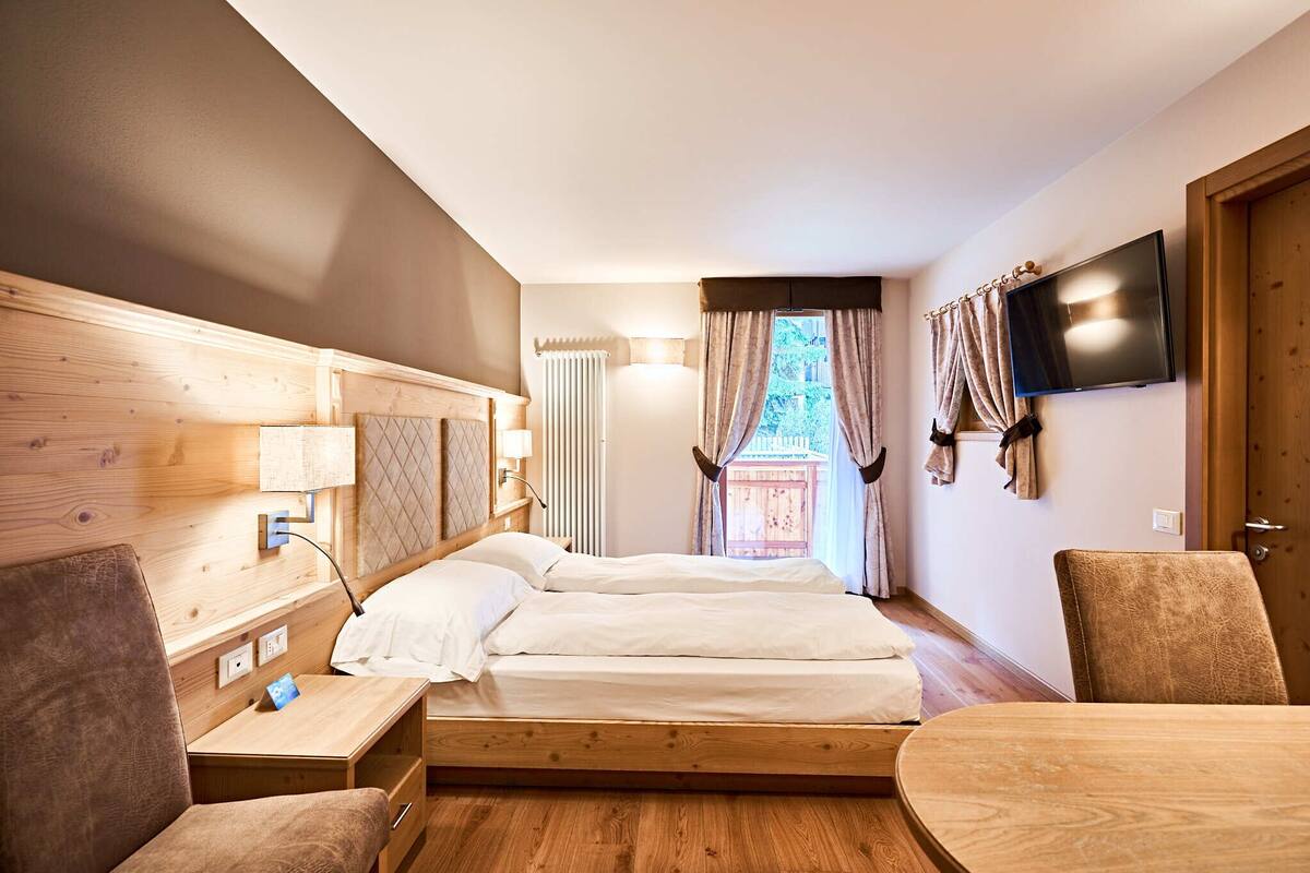 A hotel room with a wooden headboard, two beds, a TV, a table, and a view of the outdoors.