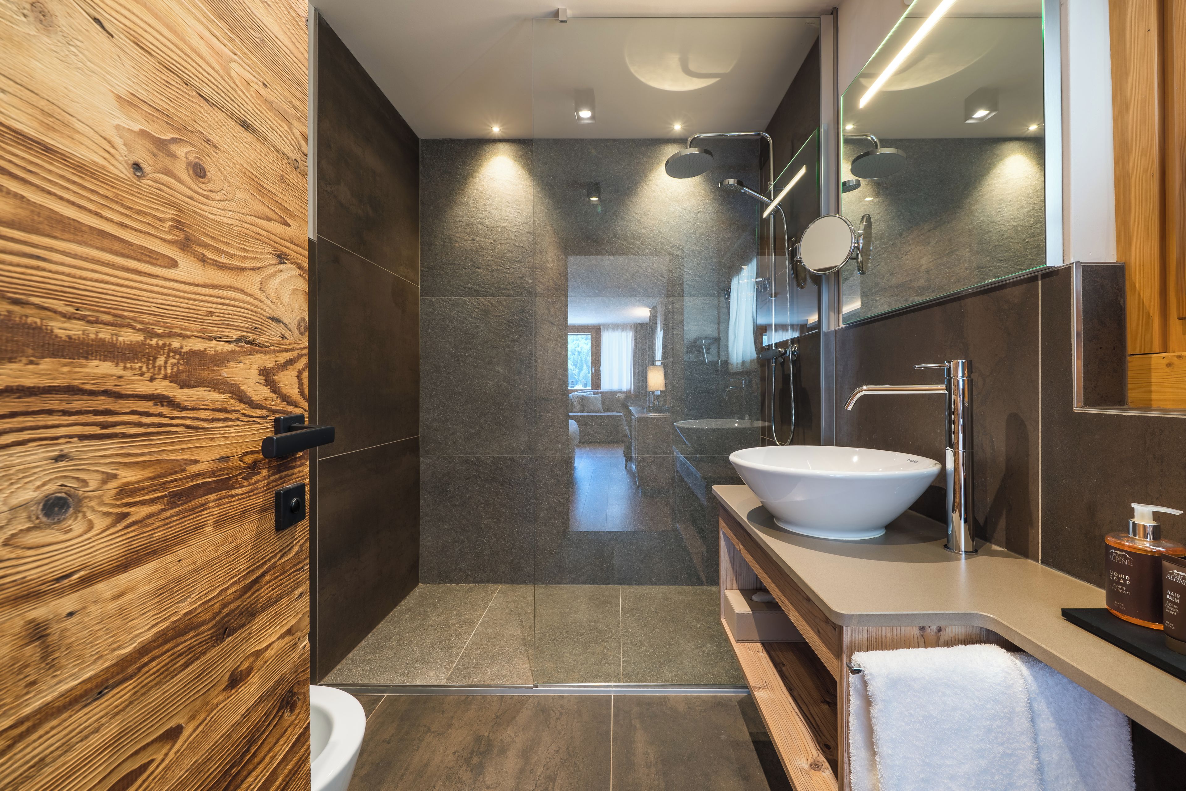 A modern bathroom with a wooden accent wall, a large mirror, a white sink, and a shower area.