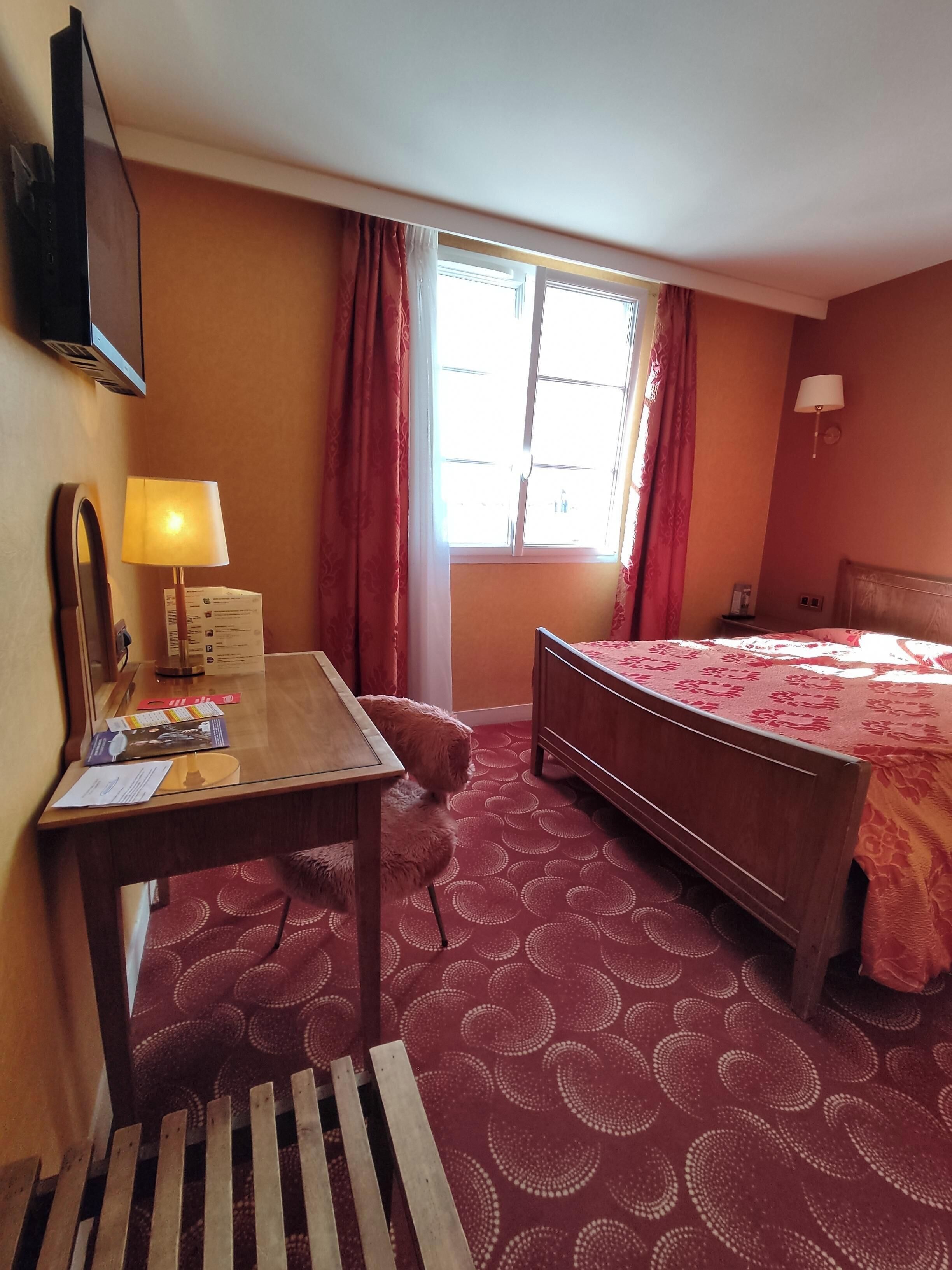 Habitación de hotel con cama de madera, un escritorio con lámpara, una ventana con cortinas y alfombra estampada.