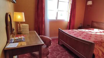 Habitación de hotel con cama de madera, un escritorio con lámpara, una ventana con cortinas y alfombra estampada.
