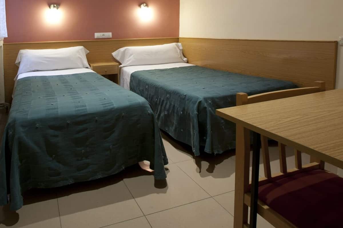 A room with two single beds, a wooden table, and two wall-mounted lights.