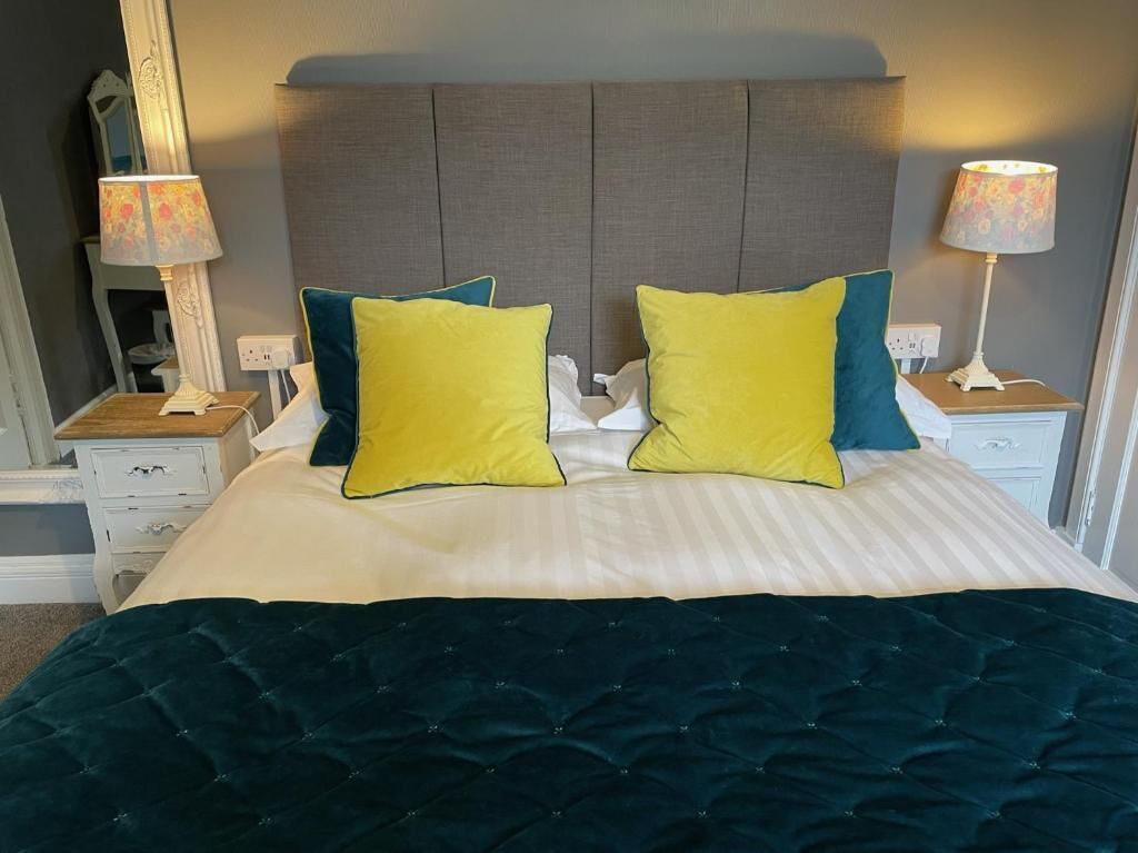 Un letto con una coperta verde scuro, due cuscini con dettagli gialli e azzurri e una testiera grigia.