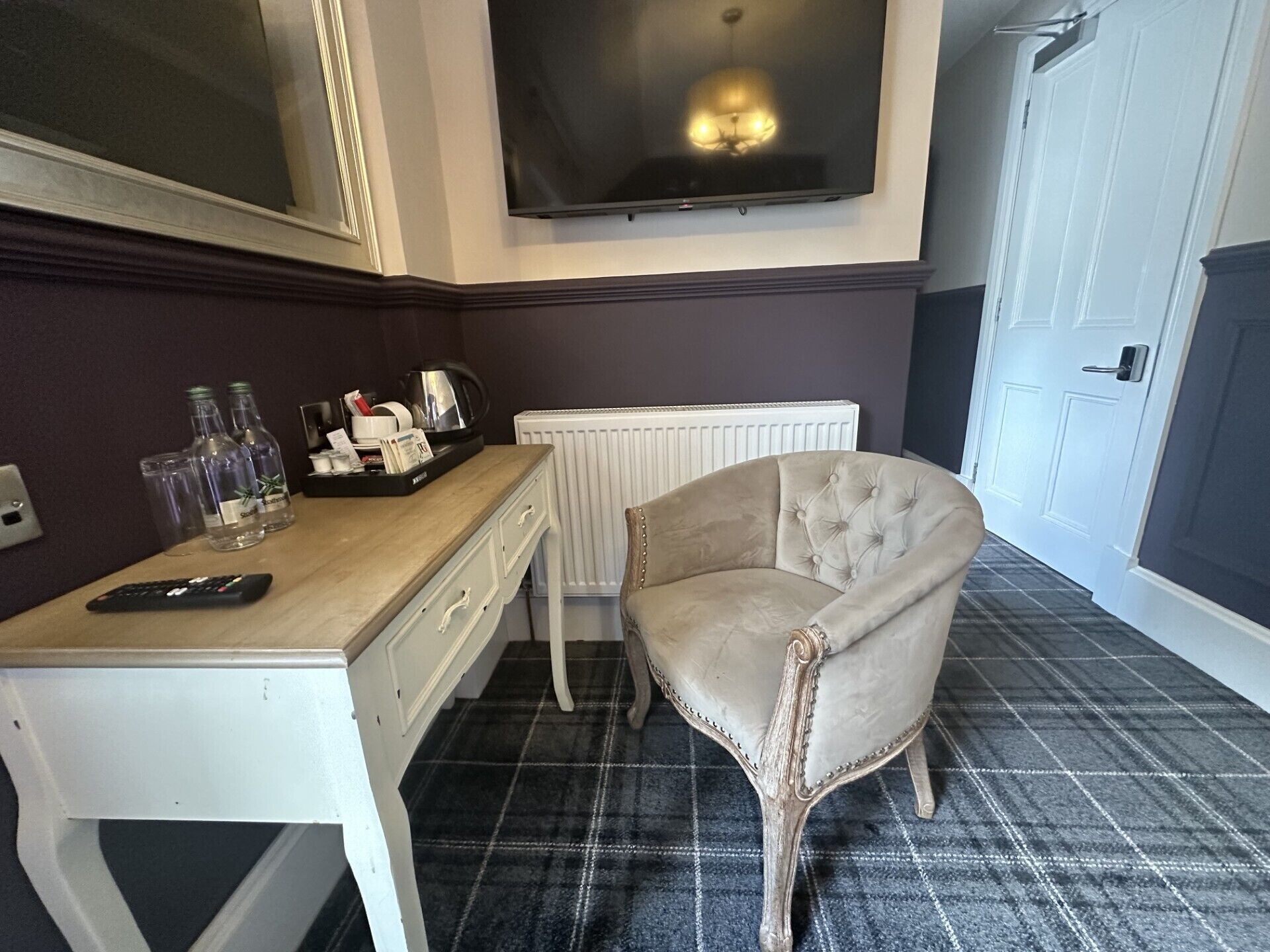 Ein Zimmer mit einem weißen Schreibtisch, einem an der Wand montierten Fernseher, einem Heizkörper und einem gepolsterten Sessel.