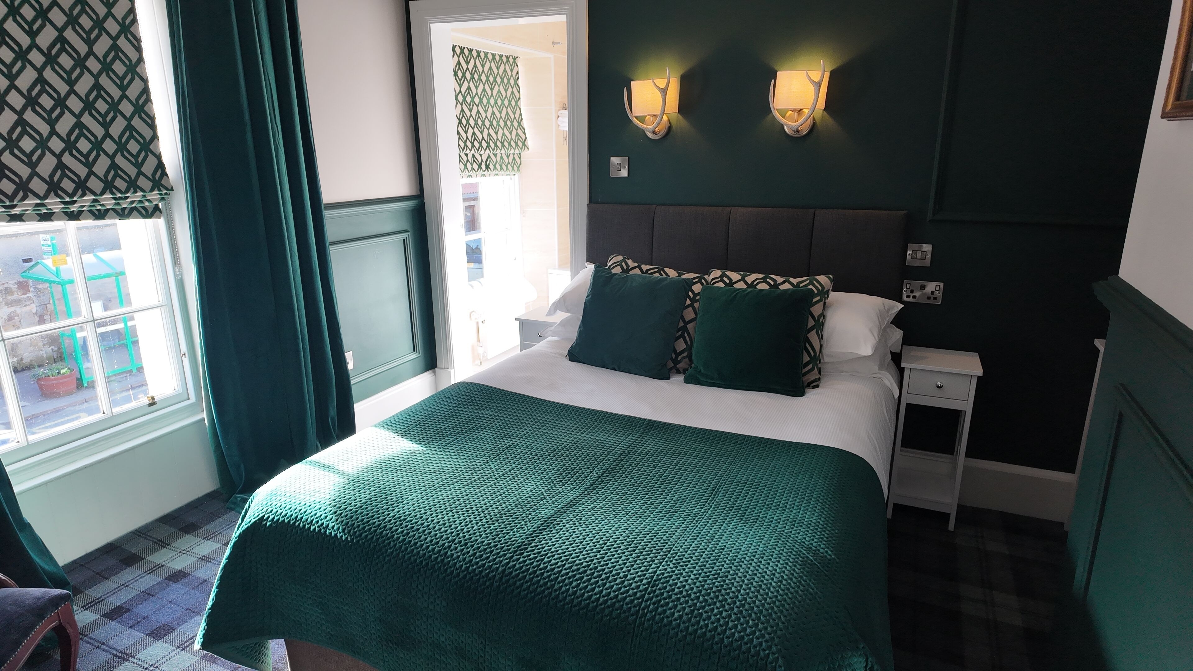 A neatly made bed with a green bedspread, two pillows, and a headboard. There is a nightstand with a lamp and a power outlet. A window with curtains is visible, and a door with a patterned curtain is also seen.
