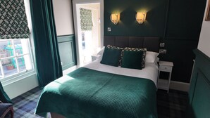 Una cama bien hecha con una colcha verde, dos almohadas y un cabecero. Se observa un mesita de noche con una lámpara y un enchufe. Se ve una ventana con cortinas, y también una puerta con un cortinaje estampado.
