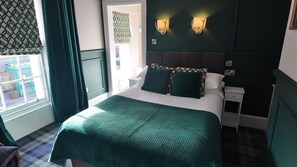 Una cama bien hecha con una colcha verde, dos almohadas y un cabecero. Se observa un mesita de noche con una lámpara y un enchufe. Se ve una ventana con cortinas, y también una puerta con un cortinaje estampado.