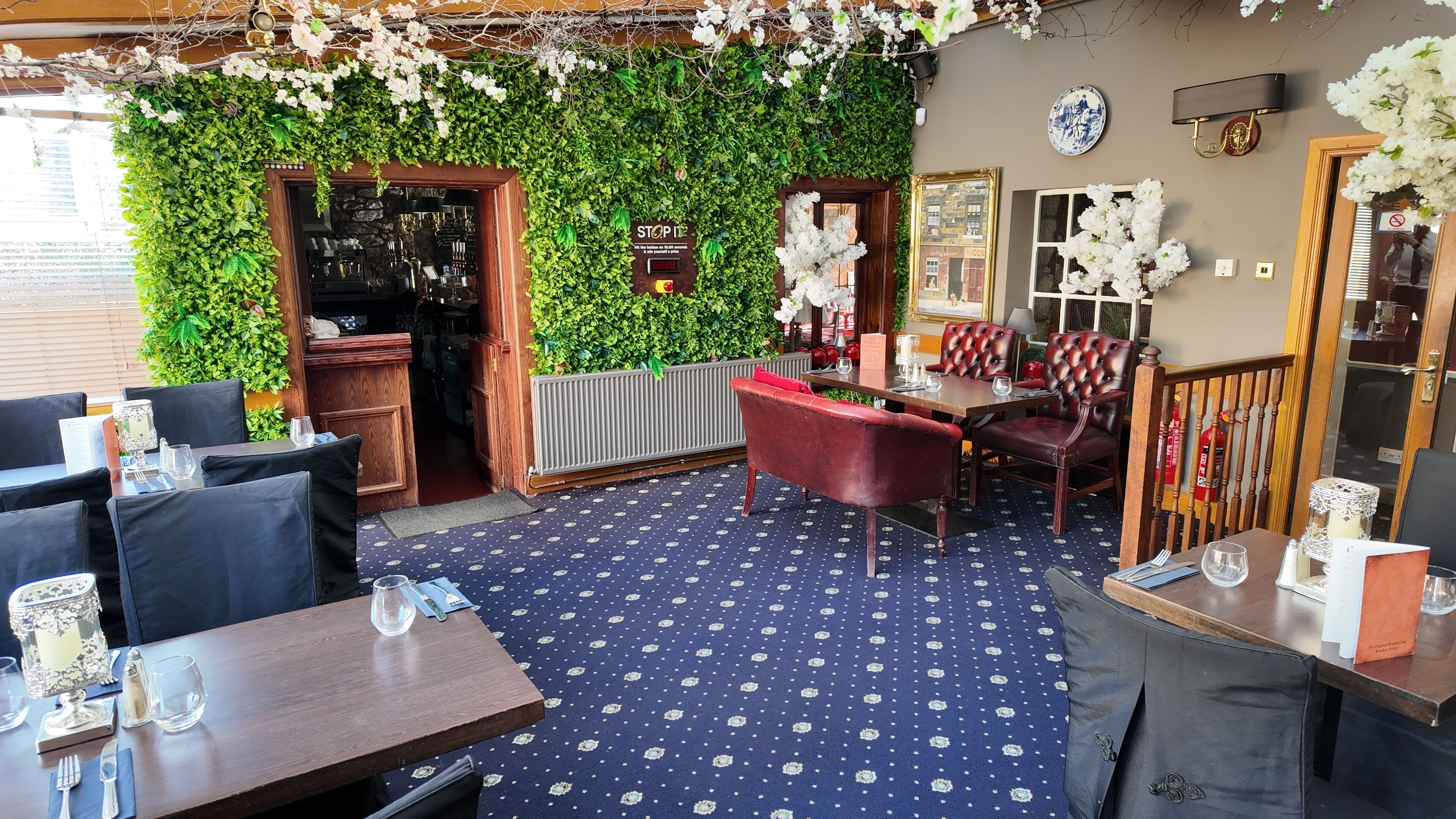 Ein Restaurantinneres mit blauem Musterteppich, Holzmöbeln und einer grünen Wand mit Kletterpflanzen.