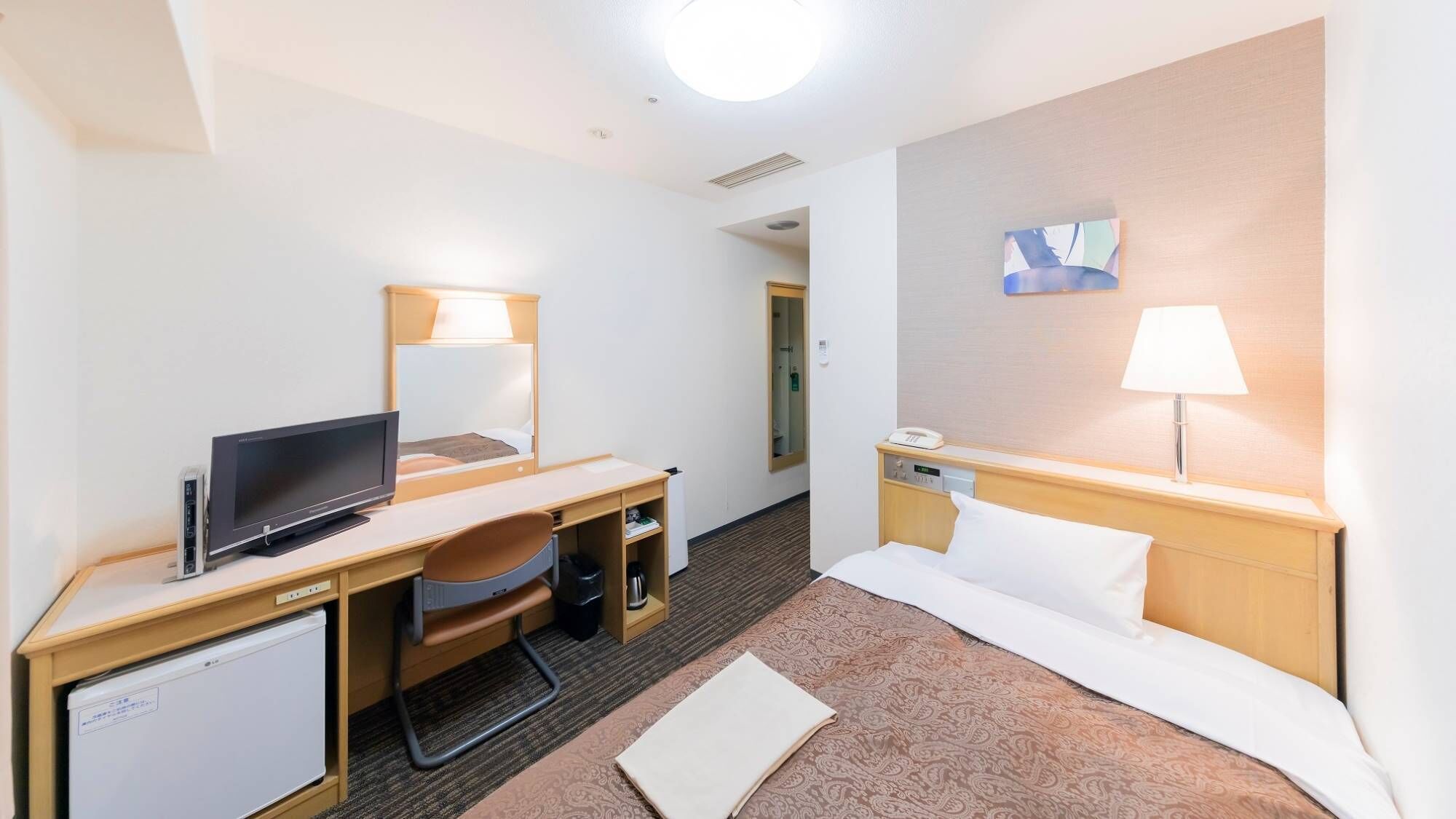 A hotel room with a bed, desk, chair, television, bedside table, lamp, and a small refrigerator.