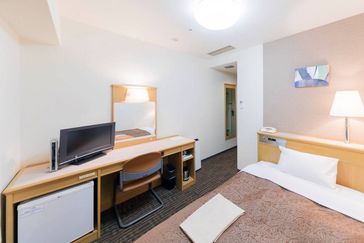 A hotel room with a bed, desk, chair, television, bedside table, lamp, and a small refrigerator.