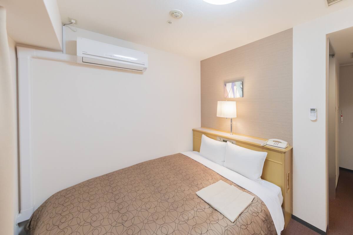 A hotel room with a bed, a bedside table with a lamp, a phone, and an air conditioning unit.