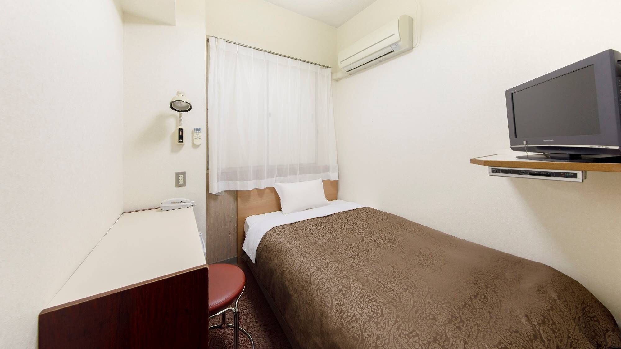 A small hotel room with a single bed, a desk with a chair, a television on a shelf, and an air conditioning unit on the wall.