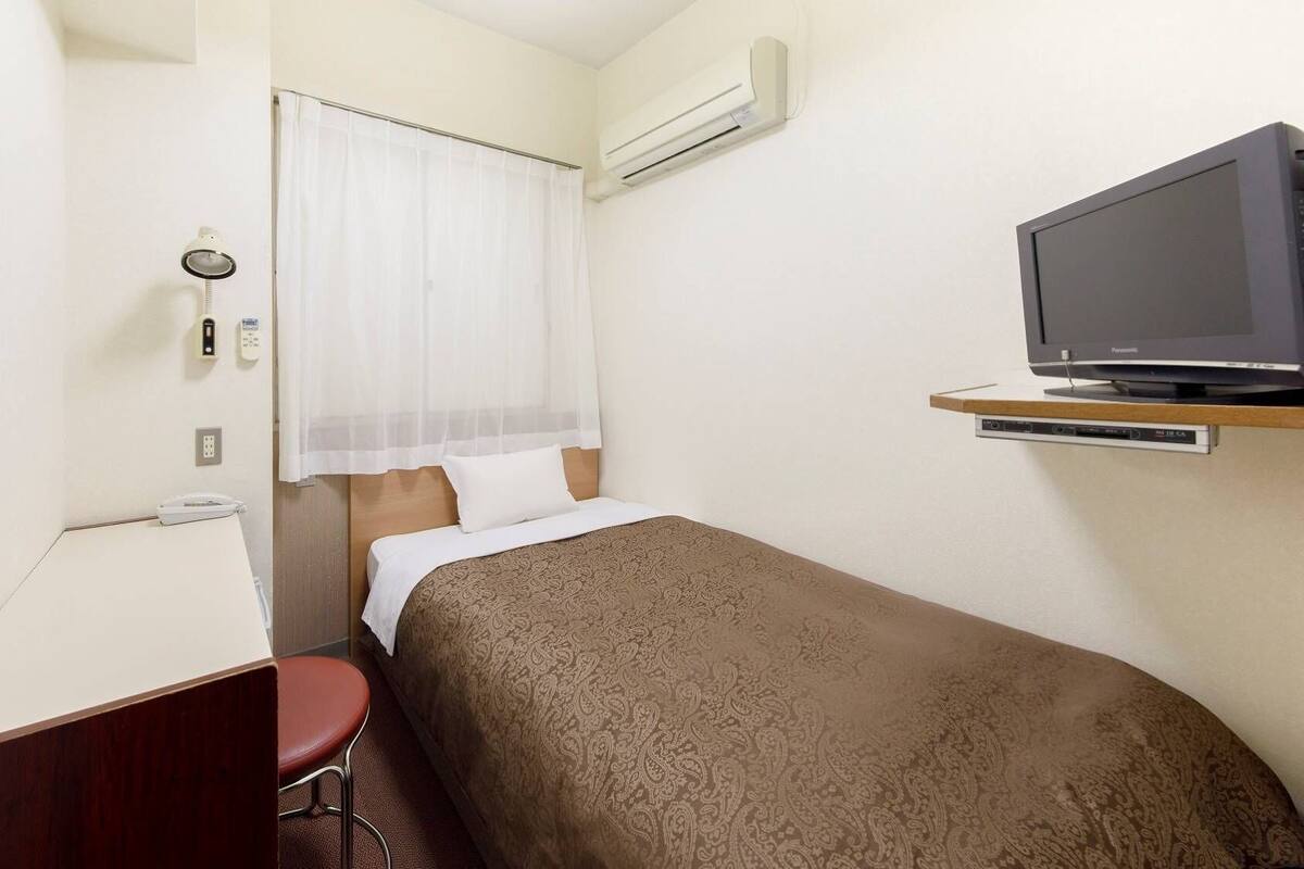 A small hotel room with a single bed, a desk with a chair, a television on a shelf, and an air conditioning unit on the wall.