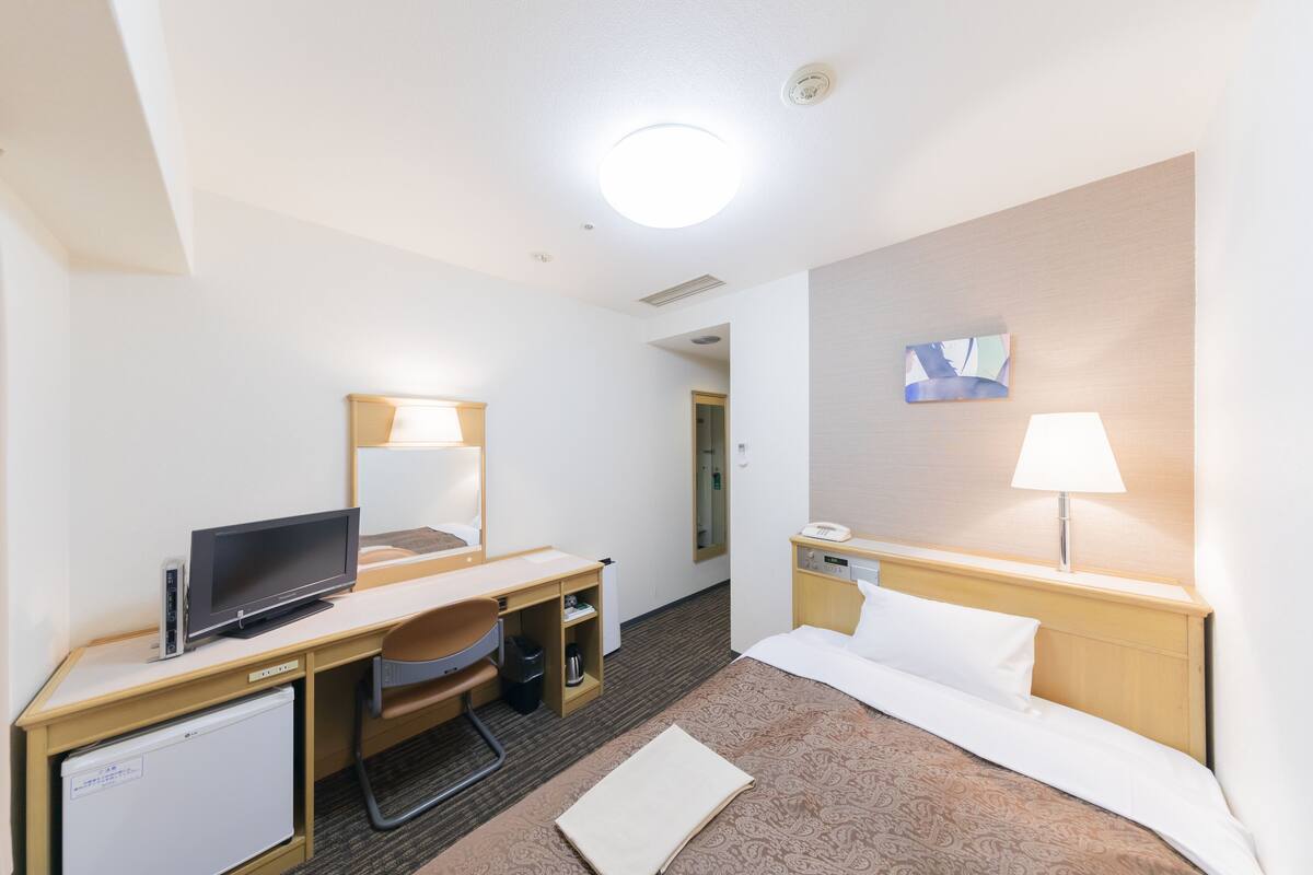 A hotel room with a bed, a desk with a computer, a TV, a mini-fridge, and a lamp.