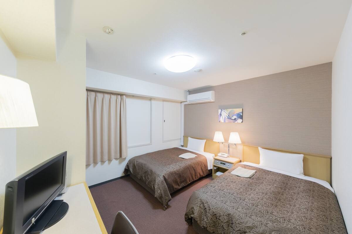A hotel room with two beds, a desk with a TV, a lamp, and an air conditioning unit on the wall.