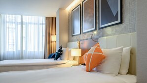 Quarto de hotel com duas camas, uma janela grande com cortinas leves, luminária fixada na parede e quadros decorativos.