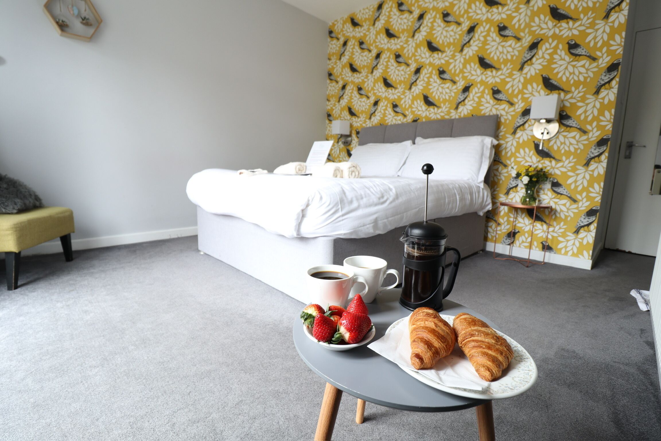 A neatly arranged bedroom with a bed, bedside tables, a coffee maker, and breakfast items including strawberries and croissants.