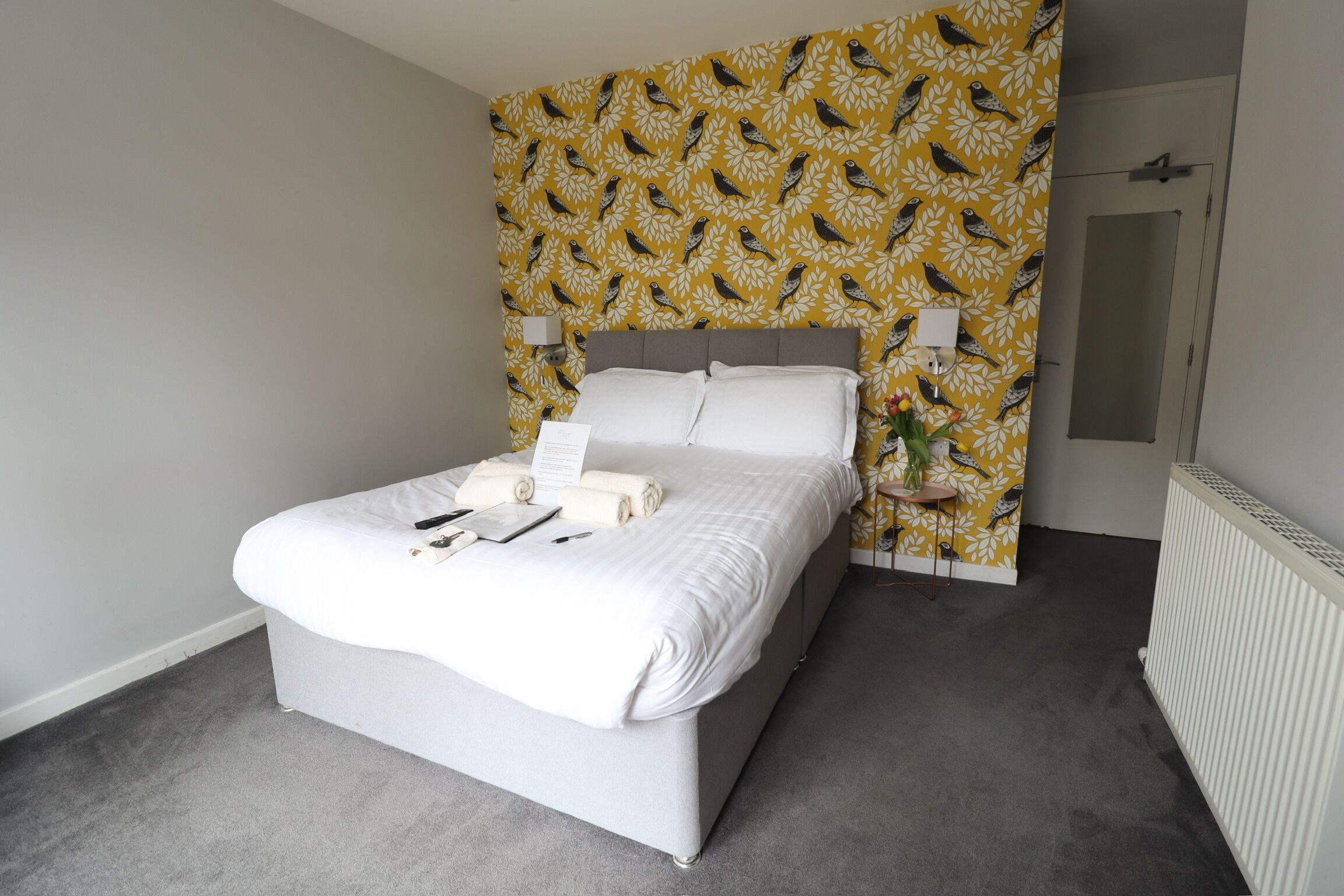 A neatly made bed with white linens and a grey headboard, a small bedside table with a lamp, and a wall with a yellow bird pattern.