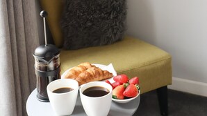 Un rincón acogedor con un sillón amarillo, una almohada gris, una mesita redonda con una cafetera, dos tazas de café, un croissant y un bol de fresas.