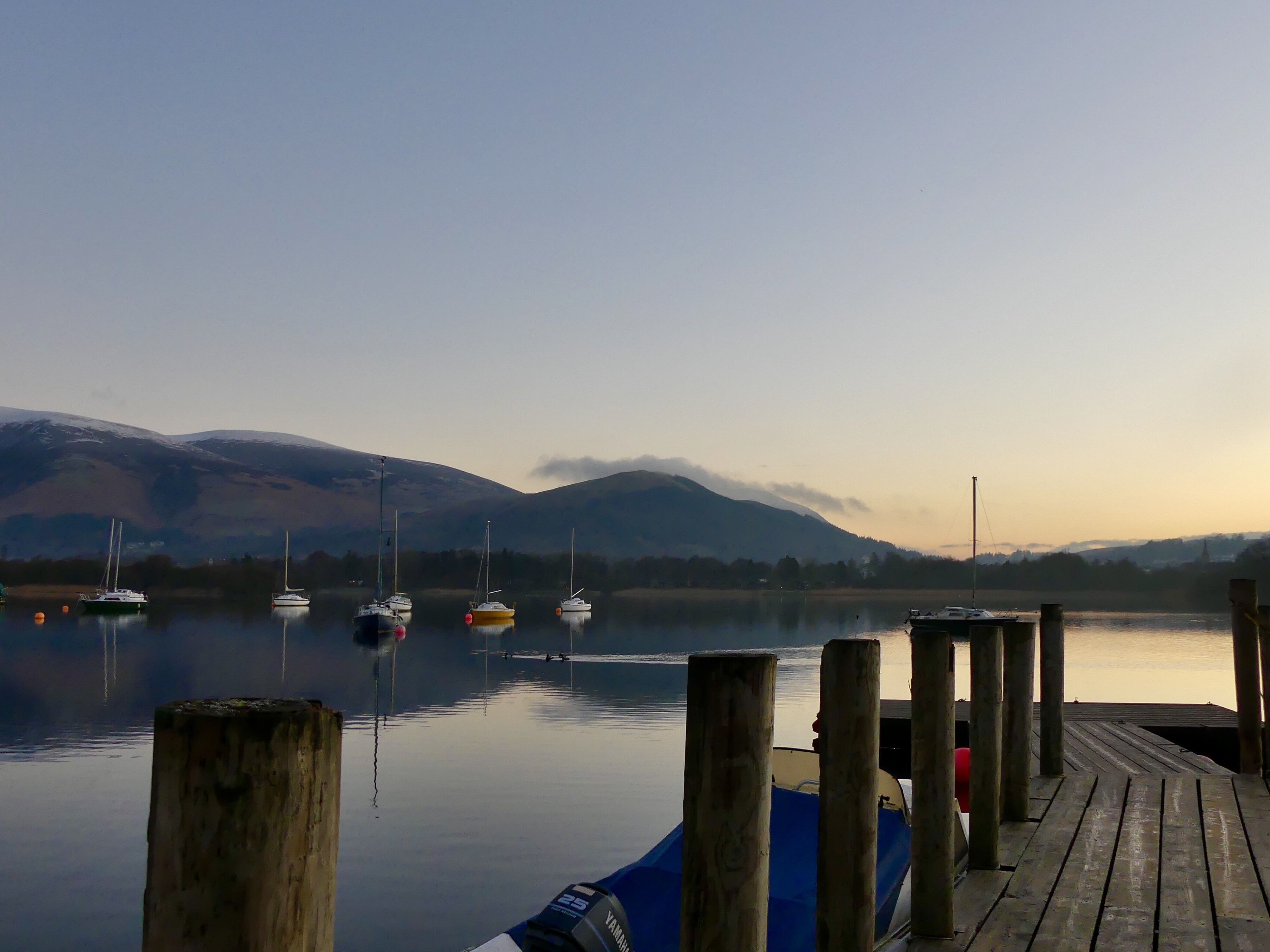 A calm lake with boats docked at wooden piers, surrounded by mountains and a clear sky.