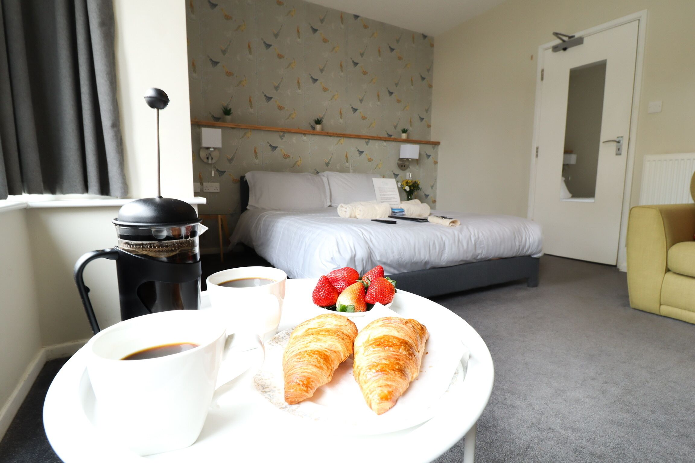 A hotel room with a bed, a coffee maker, a plate with croissants and strawberries, and a yellow armchair.