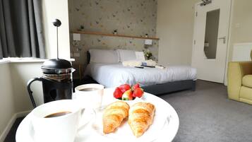 A hotel room with a bed, a coffee maker, a plate with croissants and strawberries, and a yellow armchair.