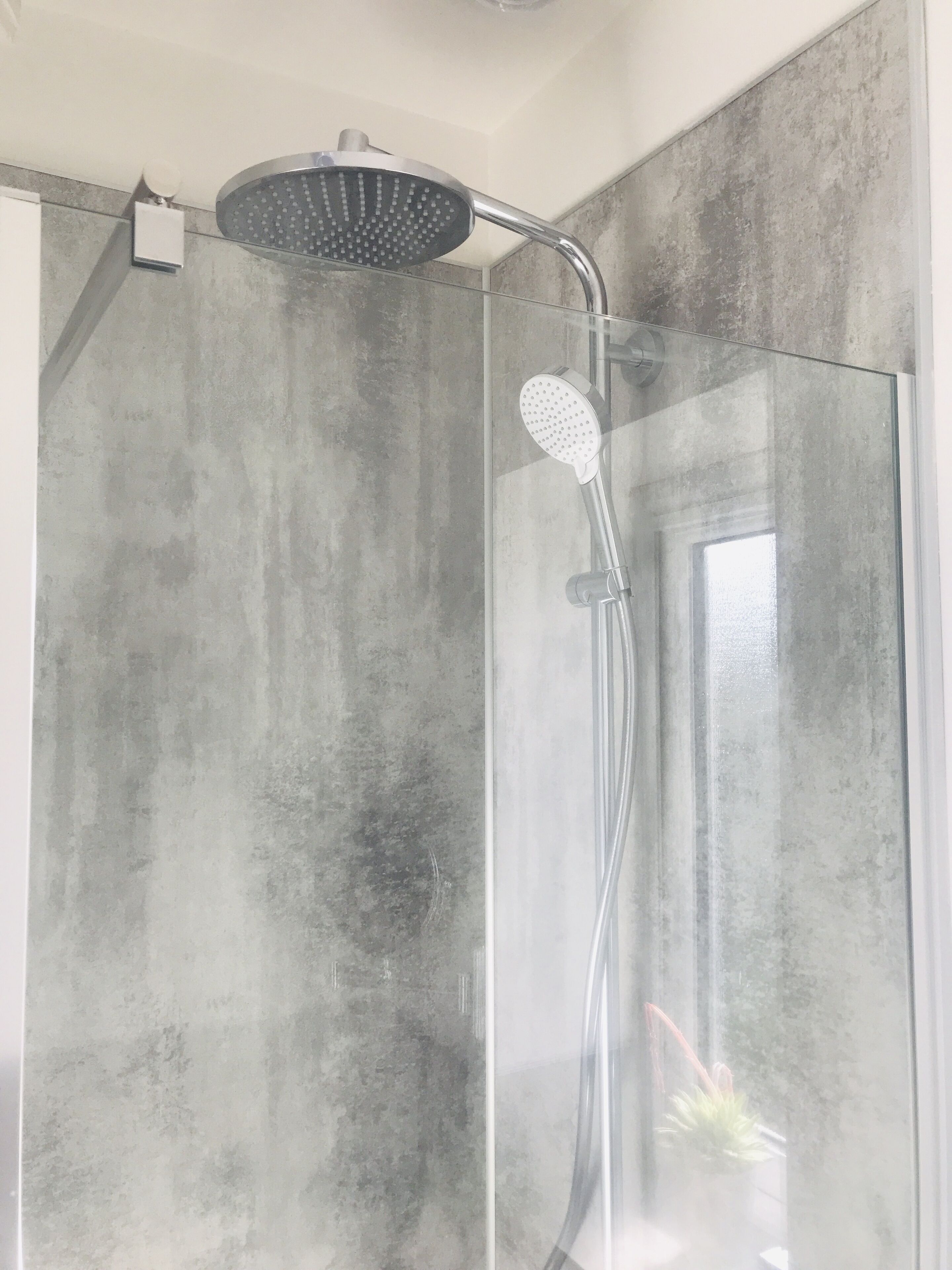 A shower with a rain showerhead and a handheld showerhead, a glass door, and a concrete wall.