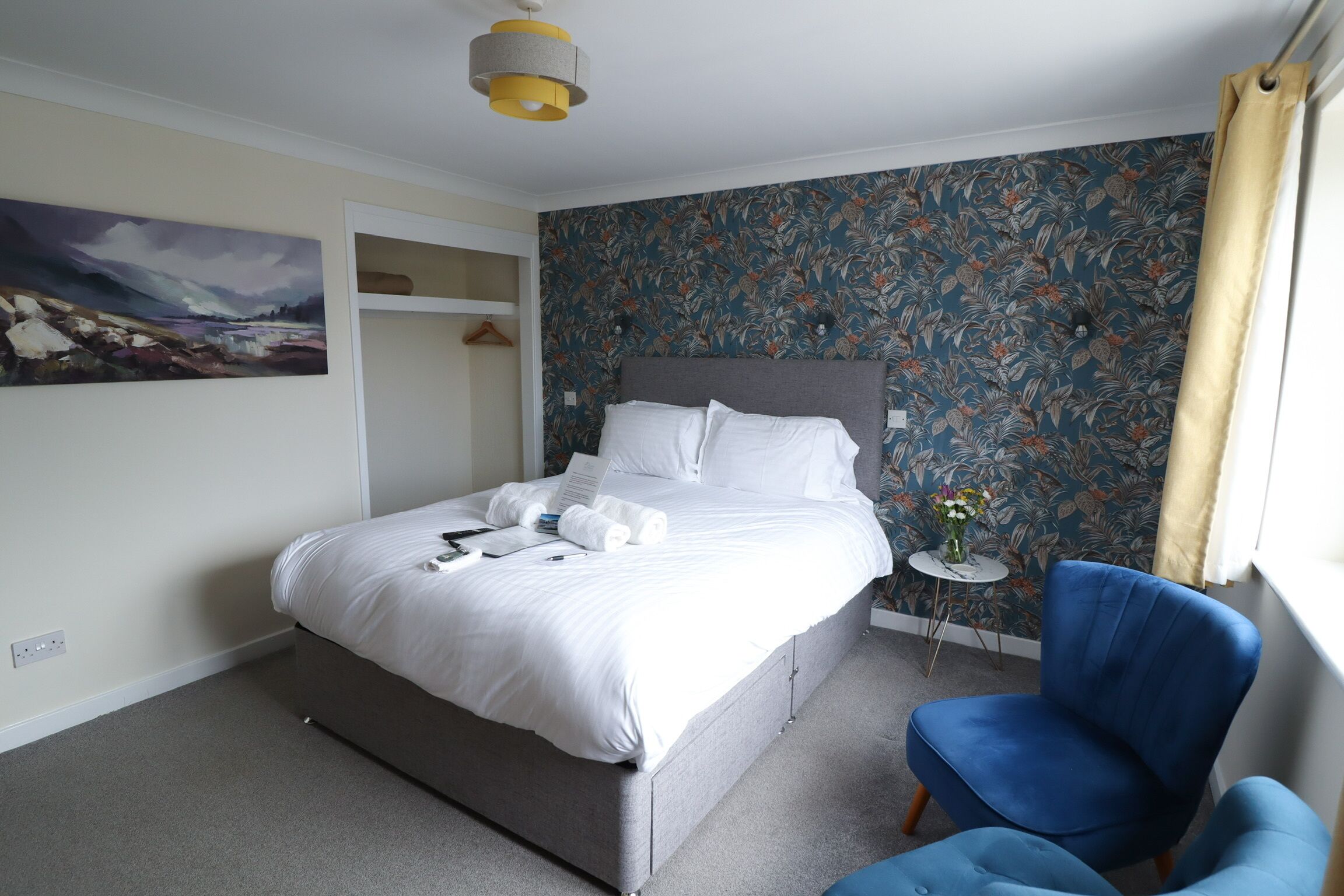 A neatly made bed with white linens, a blue armchair, and a floral wallpaper.