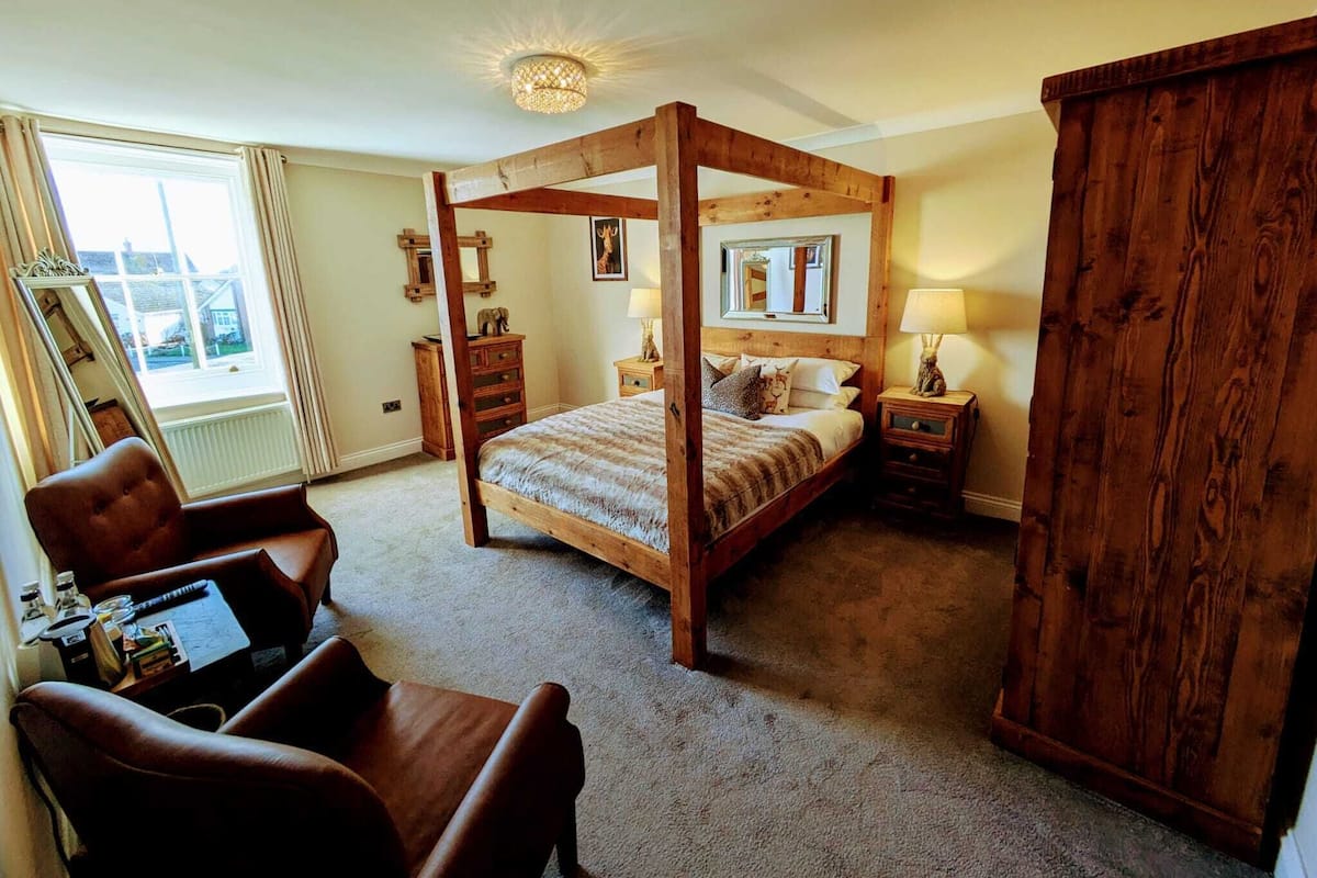 A four-poster bed with wooden headboard, a desk with a chair, a large mirror, and a window with curtains.