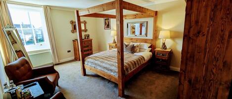 Una cama con dosel y tablero de madera, un escritorio con silla, un espejo grande y una ventana con cortinas.