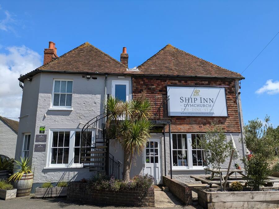 Een gebouw met twee verdiepingen, met een bord waarop staat 'Ship Inn - Dymchurch Pub, Dine, Stay'.