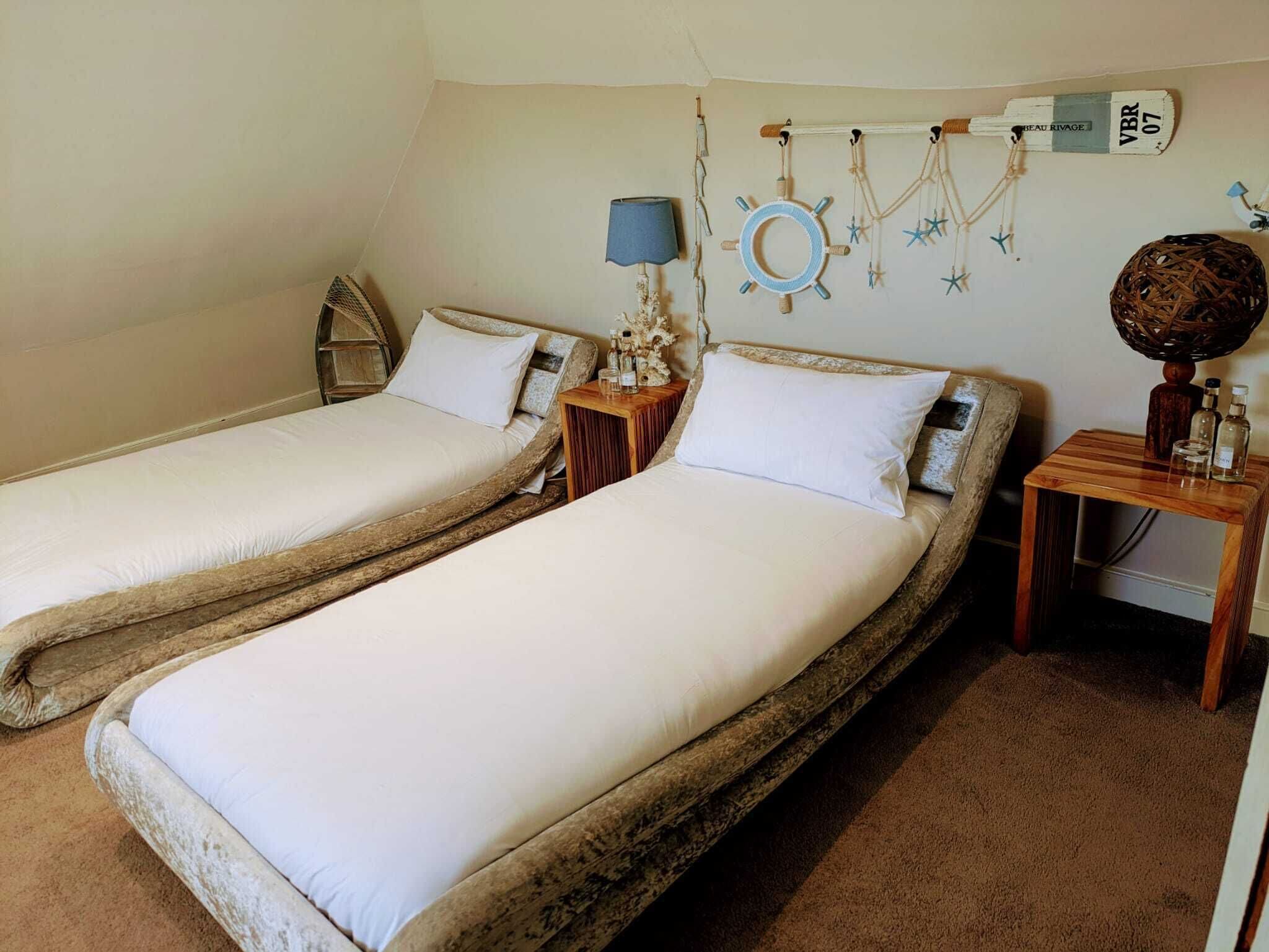 A room with two single beds, a small table with bottles, a lamp, and wall decorations including a ship's wheel and starfish.