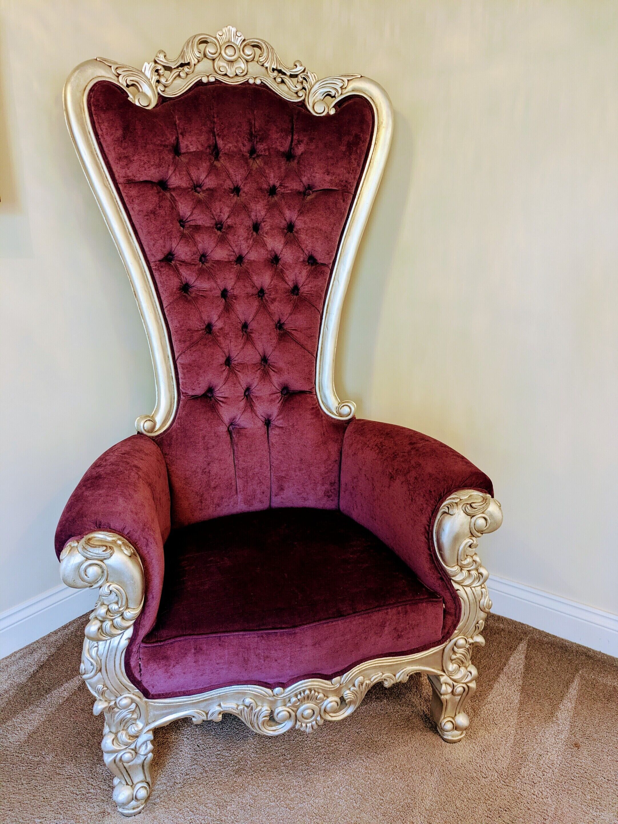 A red velvet armchair with gold ornate carvings, positioned against a plain wall.