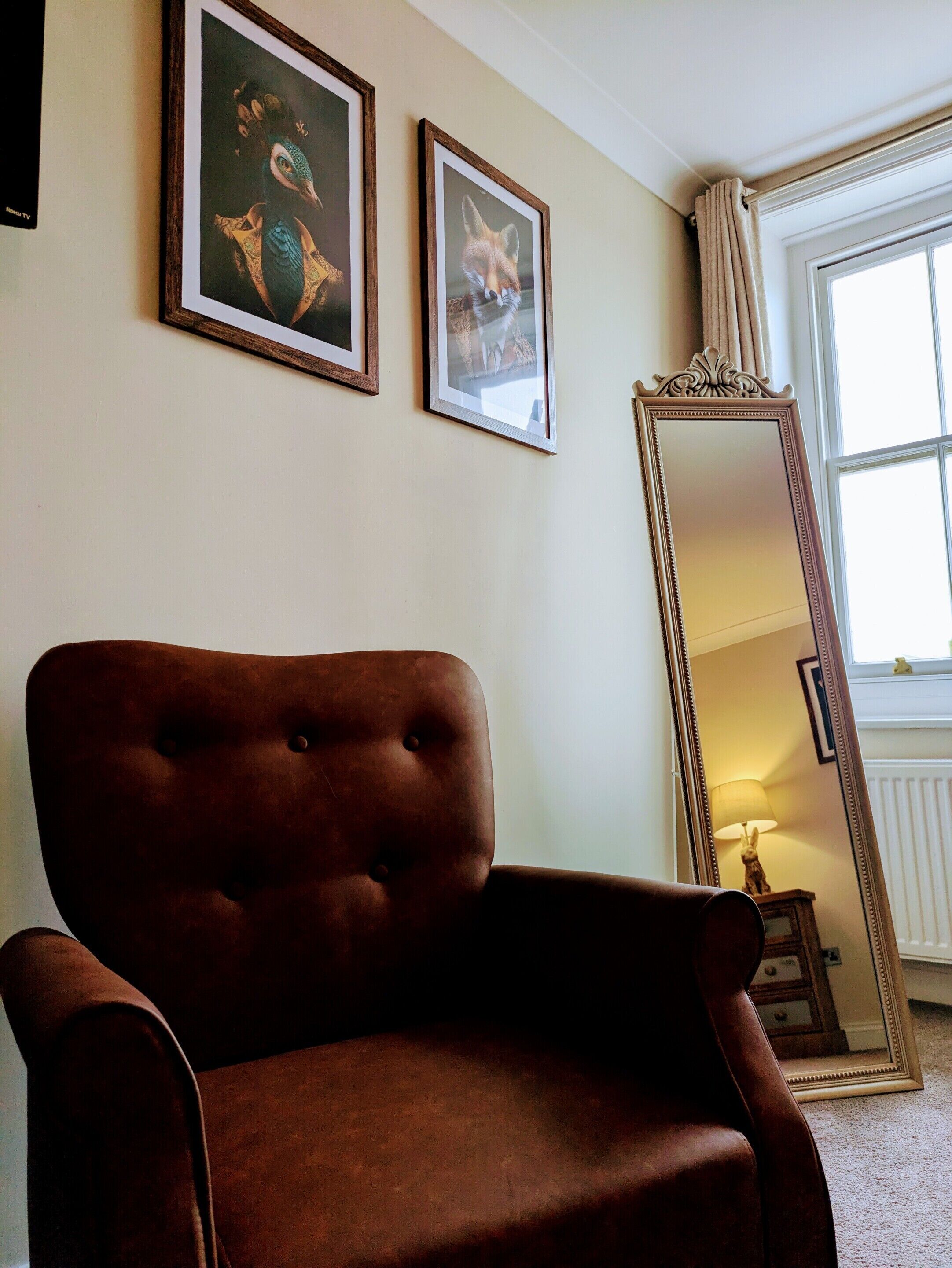 Ein Zimmer mit einem braunen Sessel, zwei gerahmten Bildern an der Wand und einem großen Spiegel, der eine Lampe und eine Statue reflektiert.