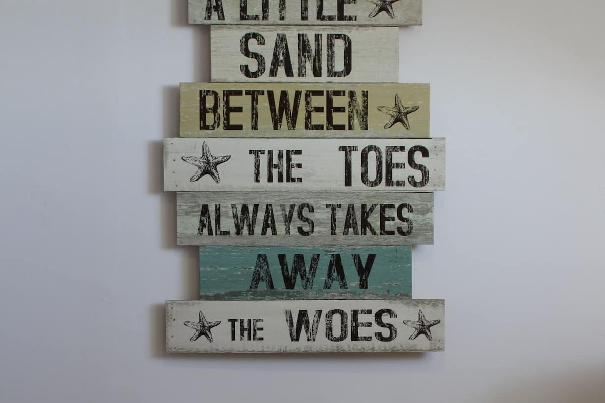 A wall with a wooden sign that reads "A LITTLE SAND BETWEEN THE TOES ALWAYS TAKES AWAY THE WOES."