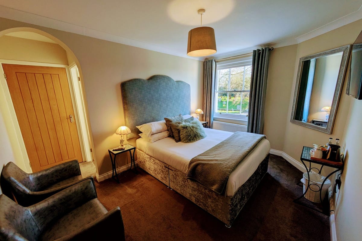 A hotel room with a large bed, a headboard, a bedside table with a lamp, a chair, a window with curtains, and a mirror on the wall.