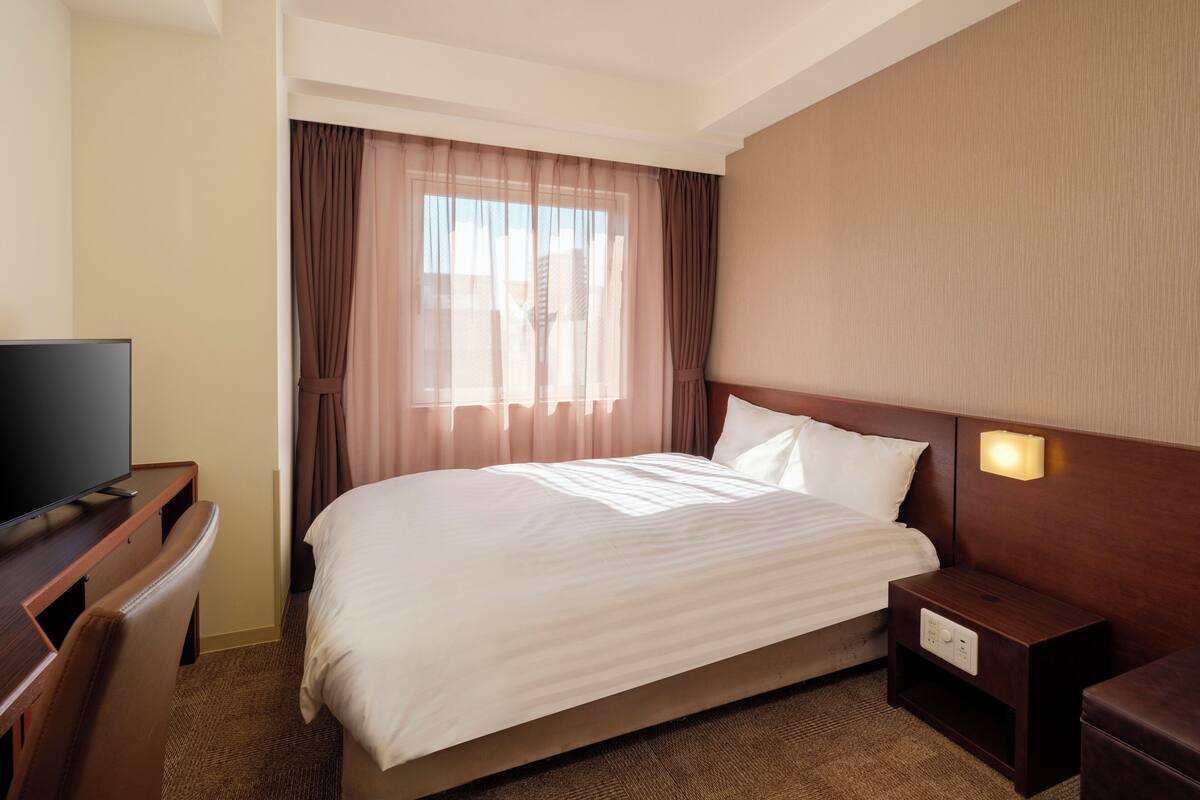 A hotel room with a bed, a TV, a nightstand, and a window with curtains.