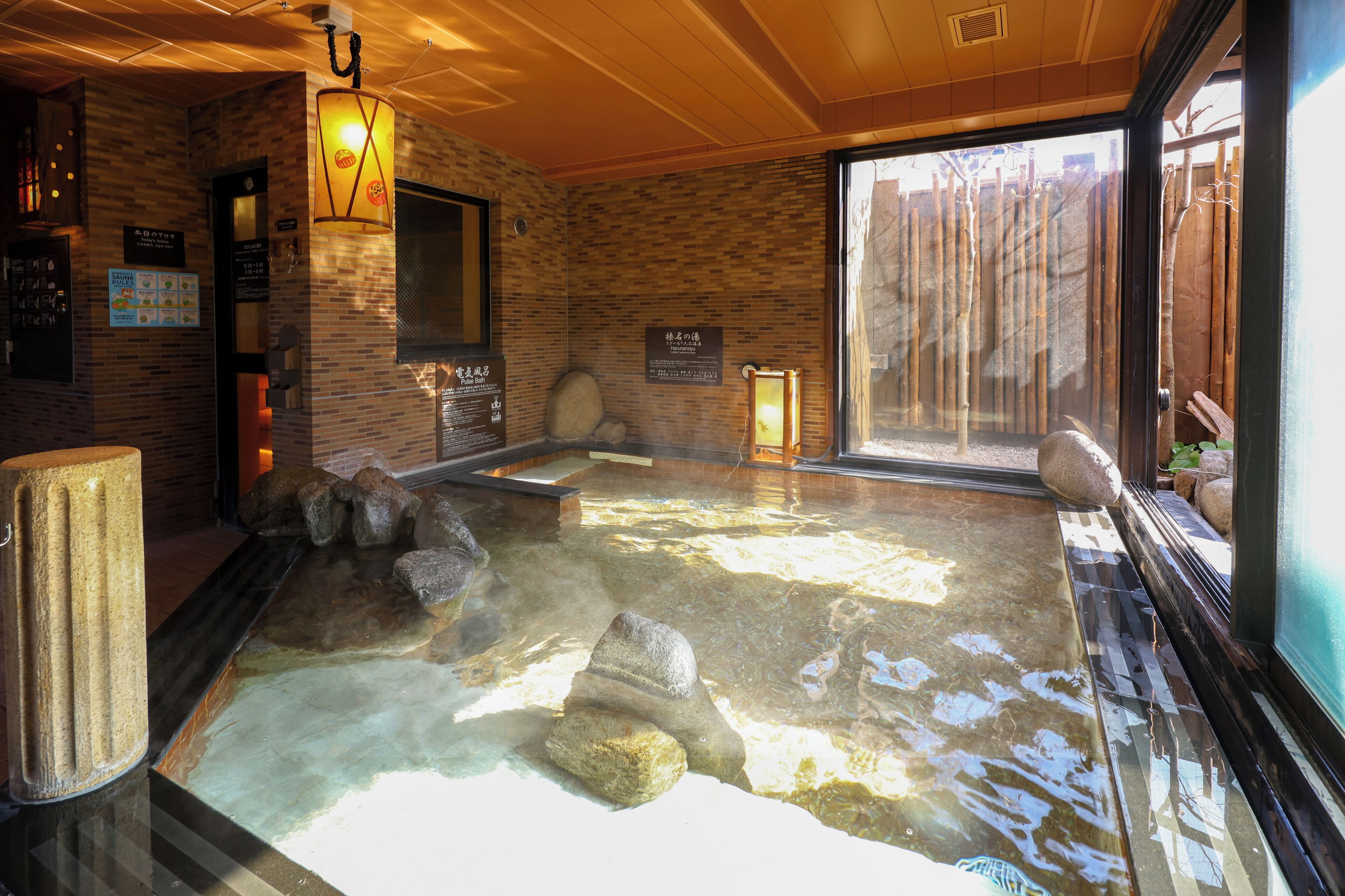 A hot spring bath with rocks, a wooden ceiling, and a bamboo fence visible through the window.