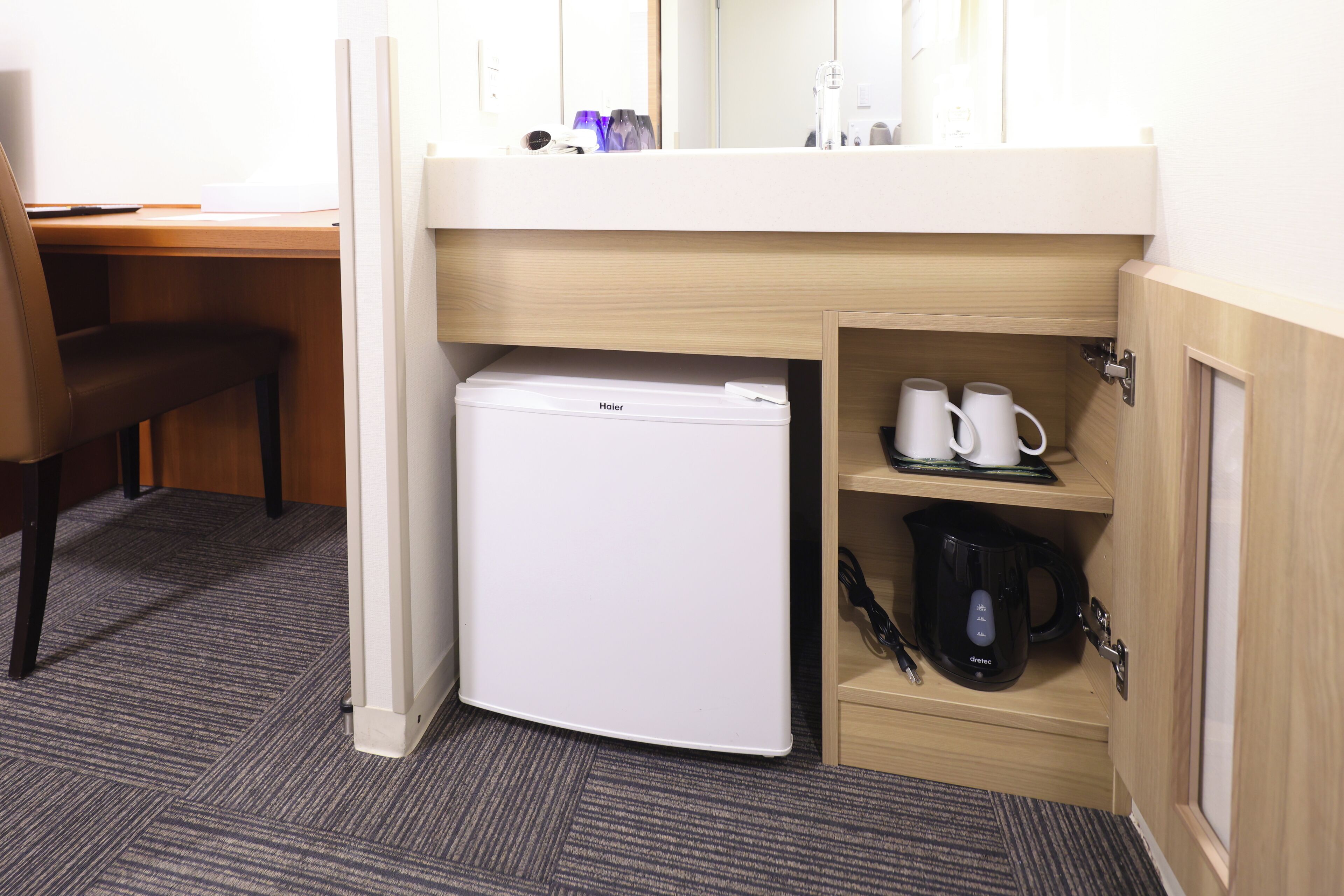 Une chambre d’hôtel de taille réduite, équipée d’un petit réfrigérateur, d’une machine à café et d’une étagère avec des tasses.