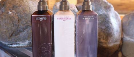 Three bottles of shampoo, conditioner, and body soap on a metal surface with a brick wall and large rocks in the background.