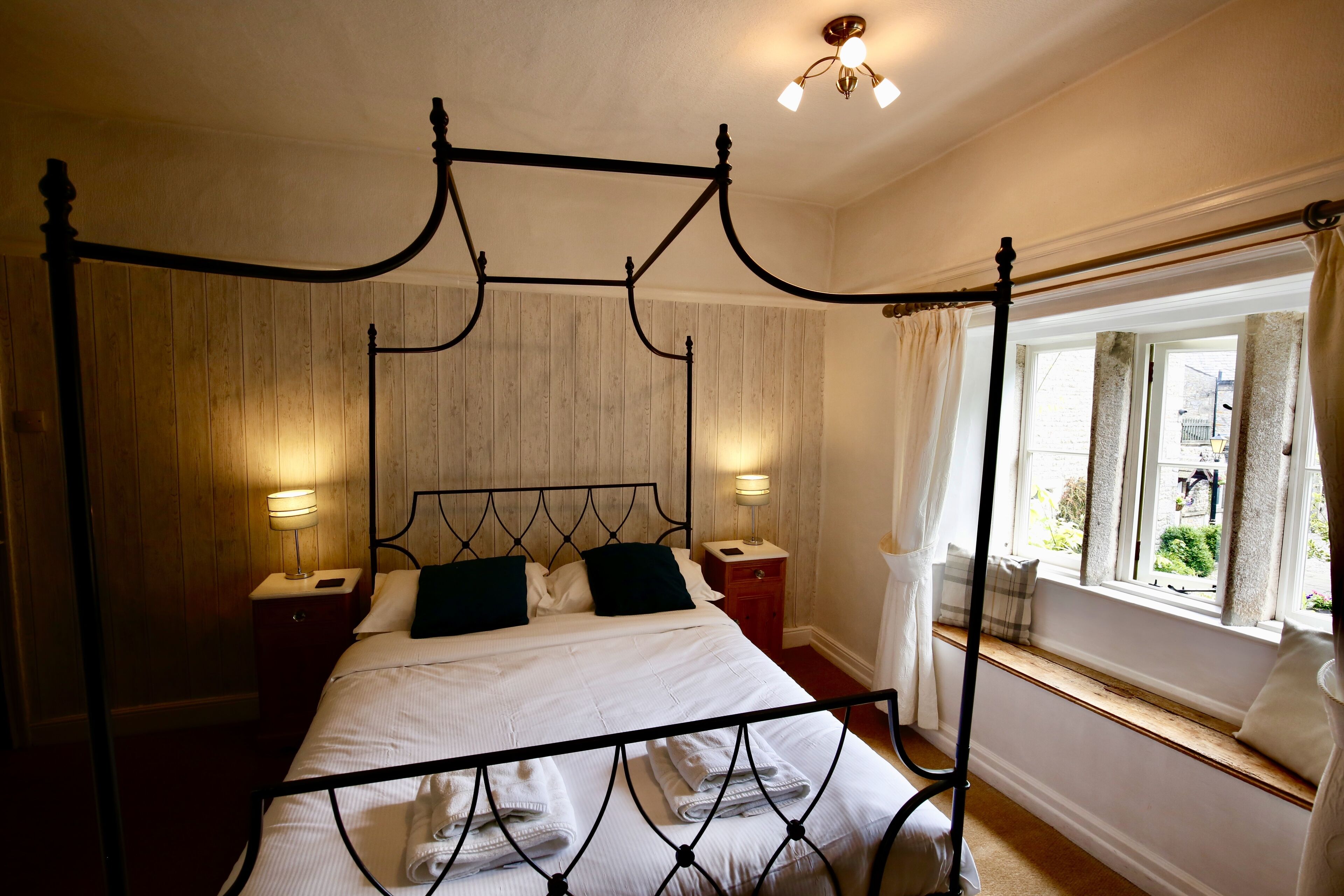 Una cama con dosel, cabecera de madera, dos mesitas de noche con lámparas y una ventana con cortinas.
