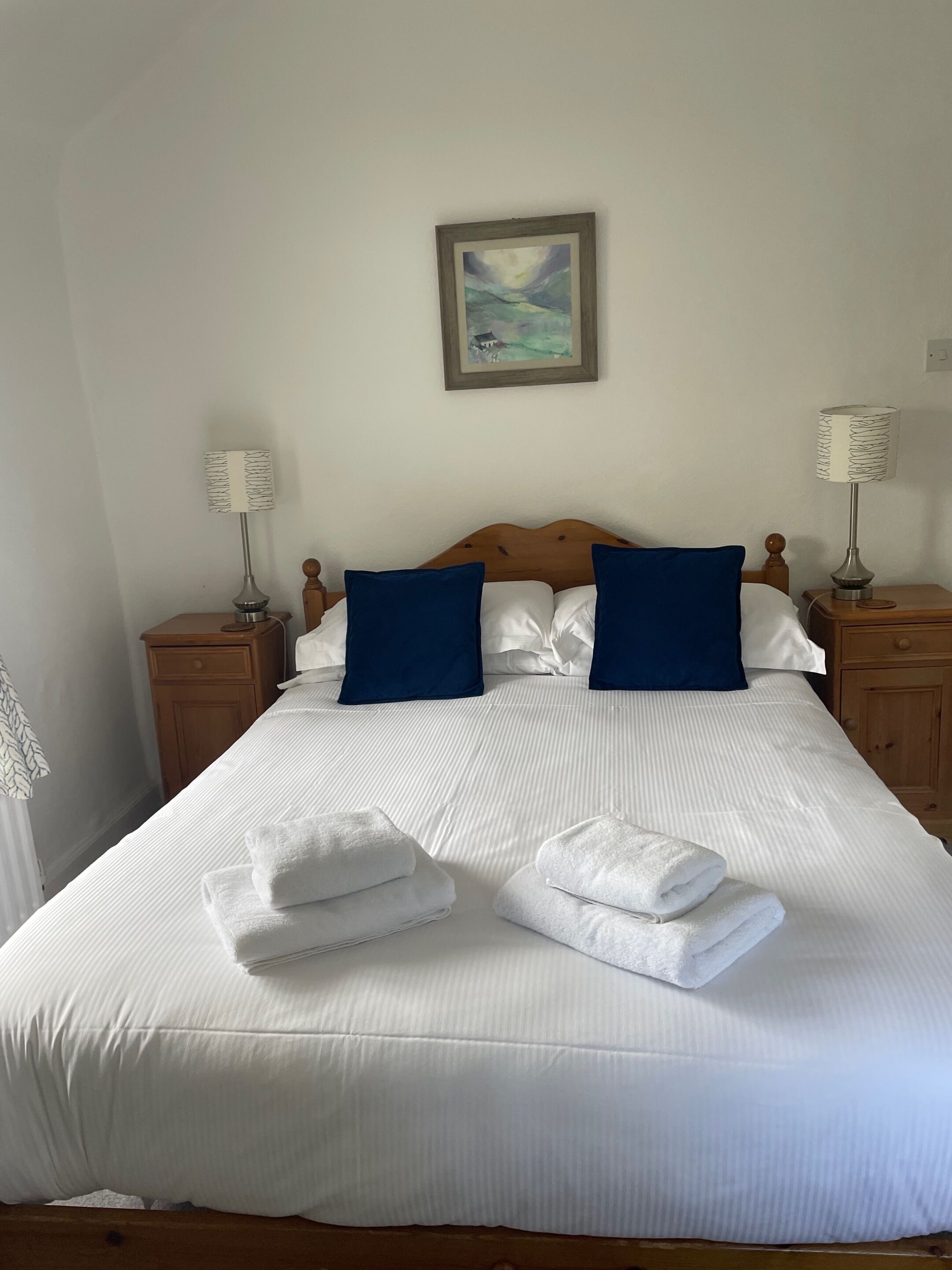 A neatly made bed with white linens, two blue pillows, and folded towels on the bed.