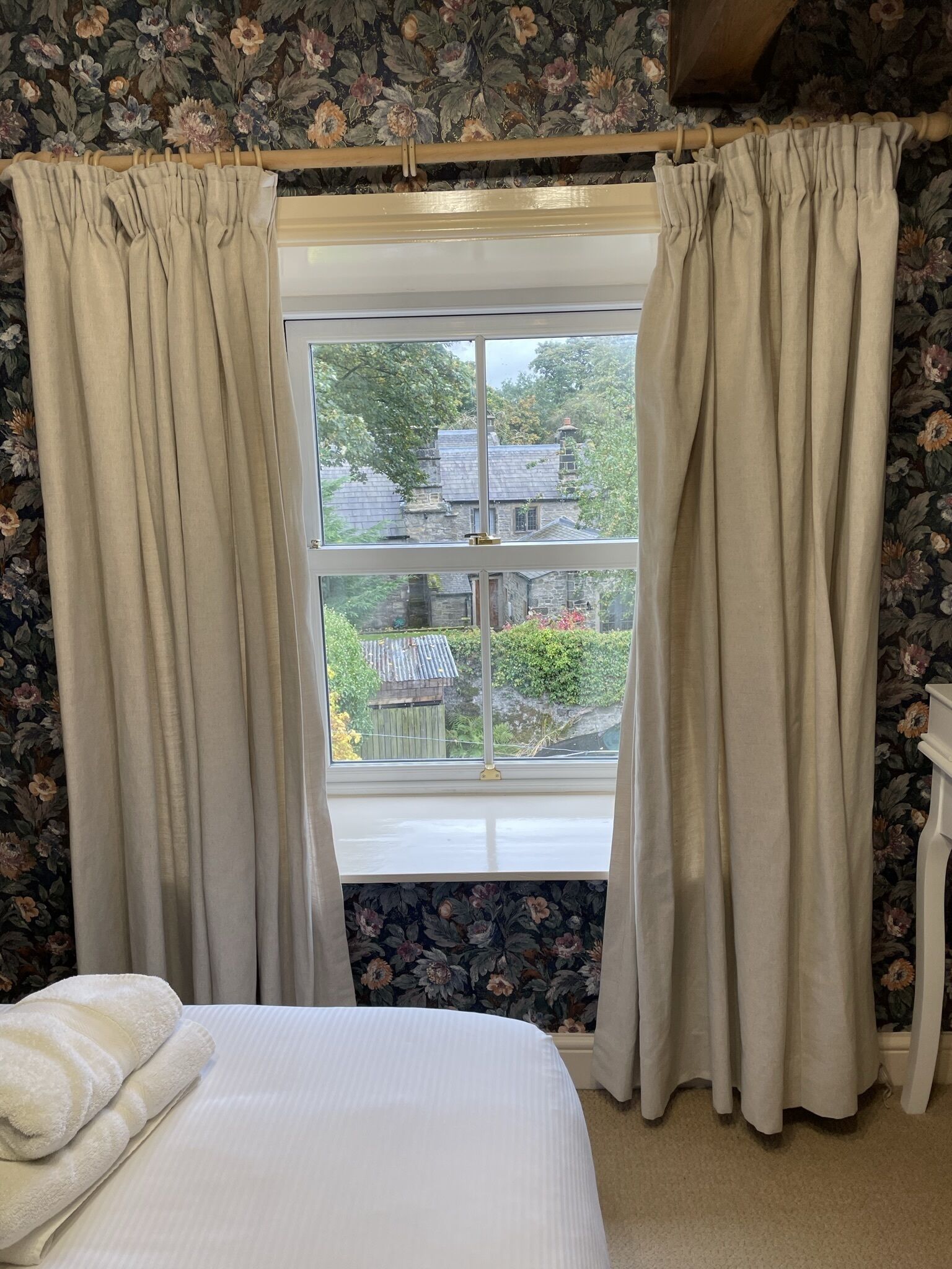A bedroom with a bed, beige curtains, floral wallpaper, and a window with a view of a garden and buildings.