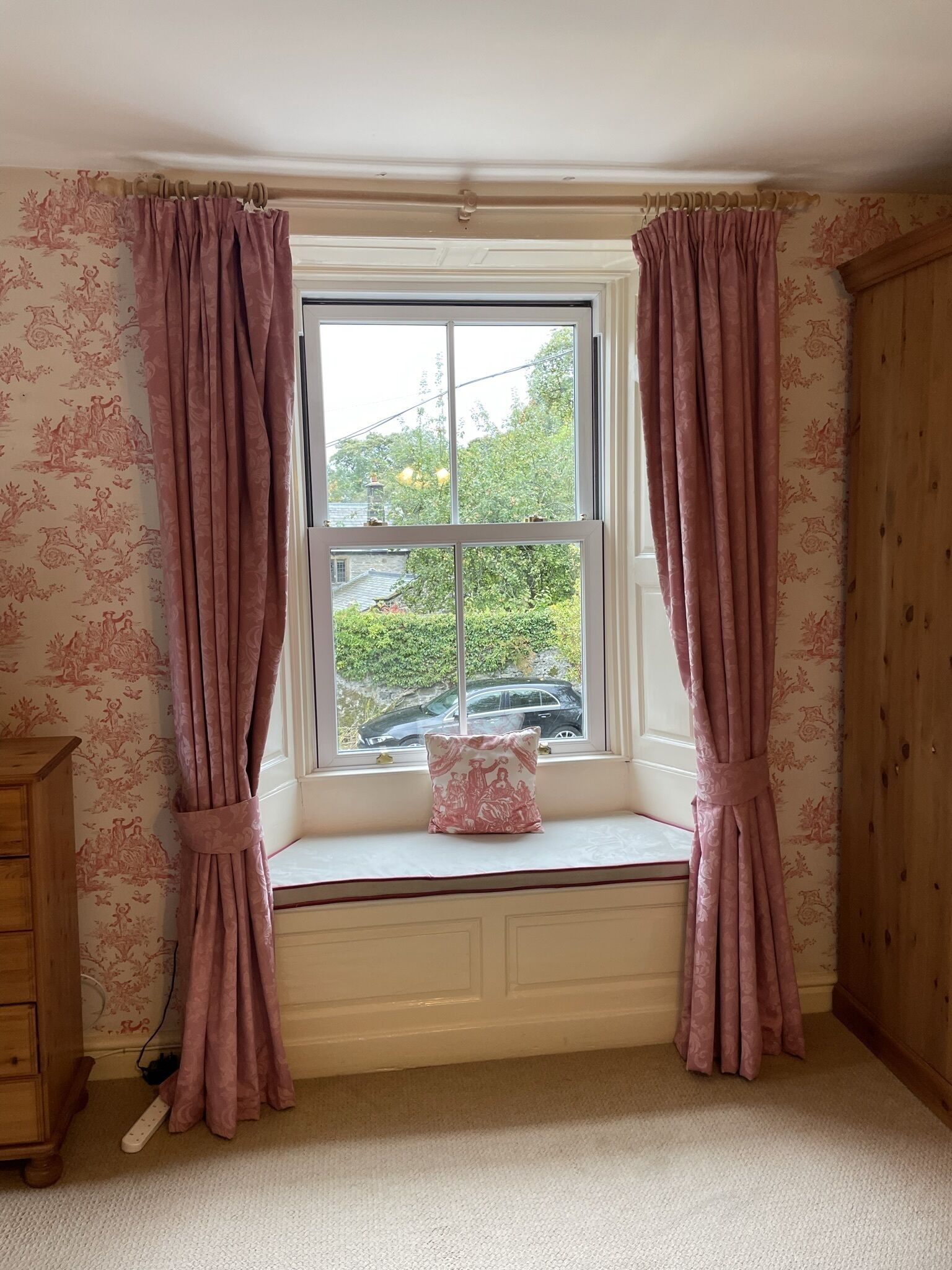 Una habitación con asiento junto a la ventana, cortinas, un armario de madera y un papel tapiz estampado.