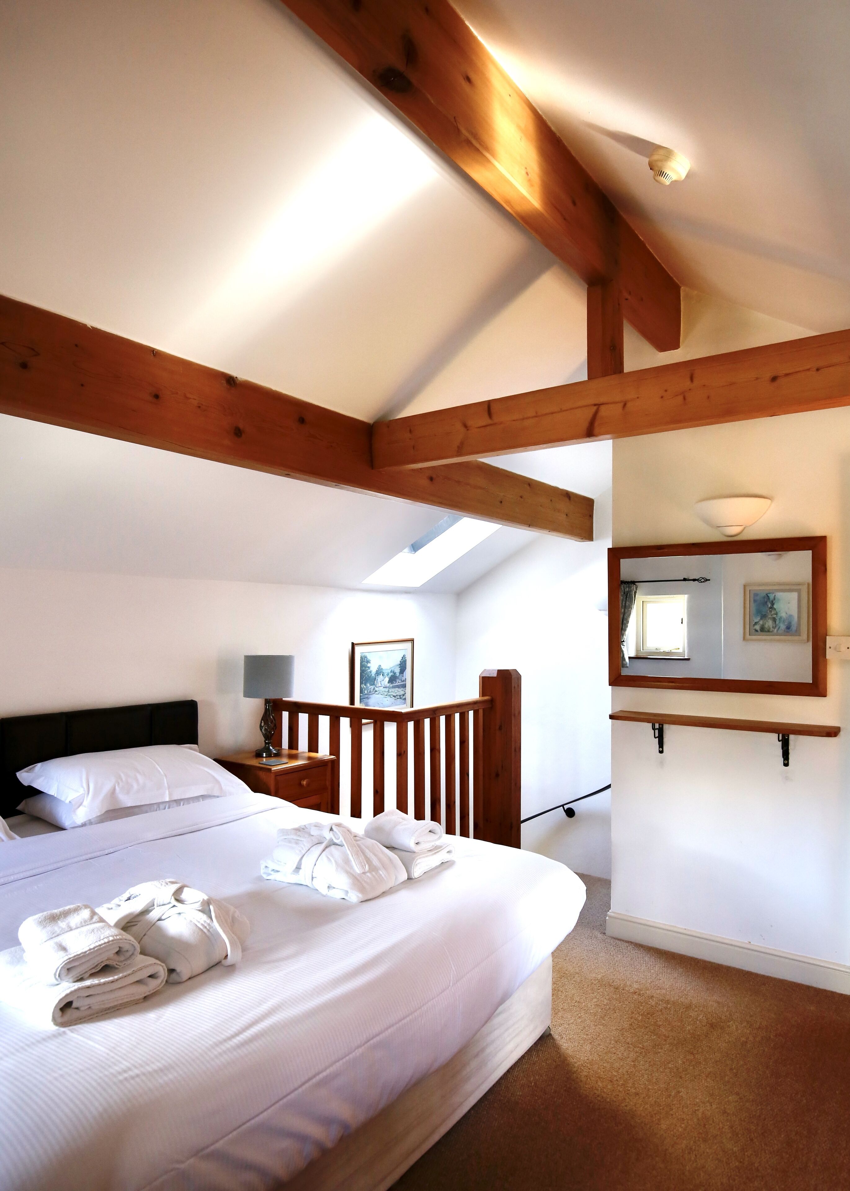 A bedroom with a large bed, wooden beams, a skylight, a bedside table with a lamp, and a framed picture on the wall.