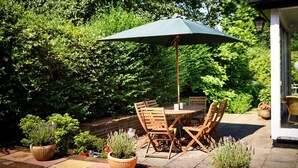 Um pátio externo com mesa e cadeiras sob um grande guarda-sol, cercado por plantas em vasos e uma parede de tijolos.