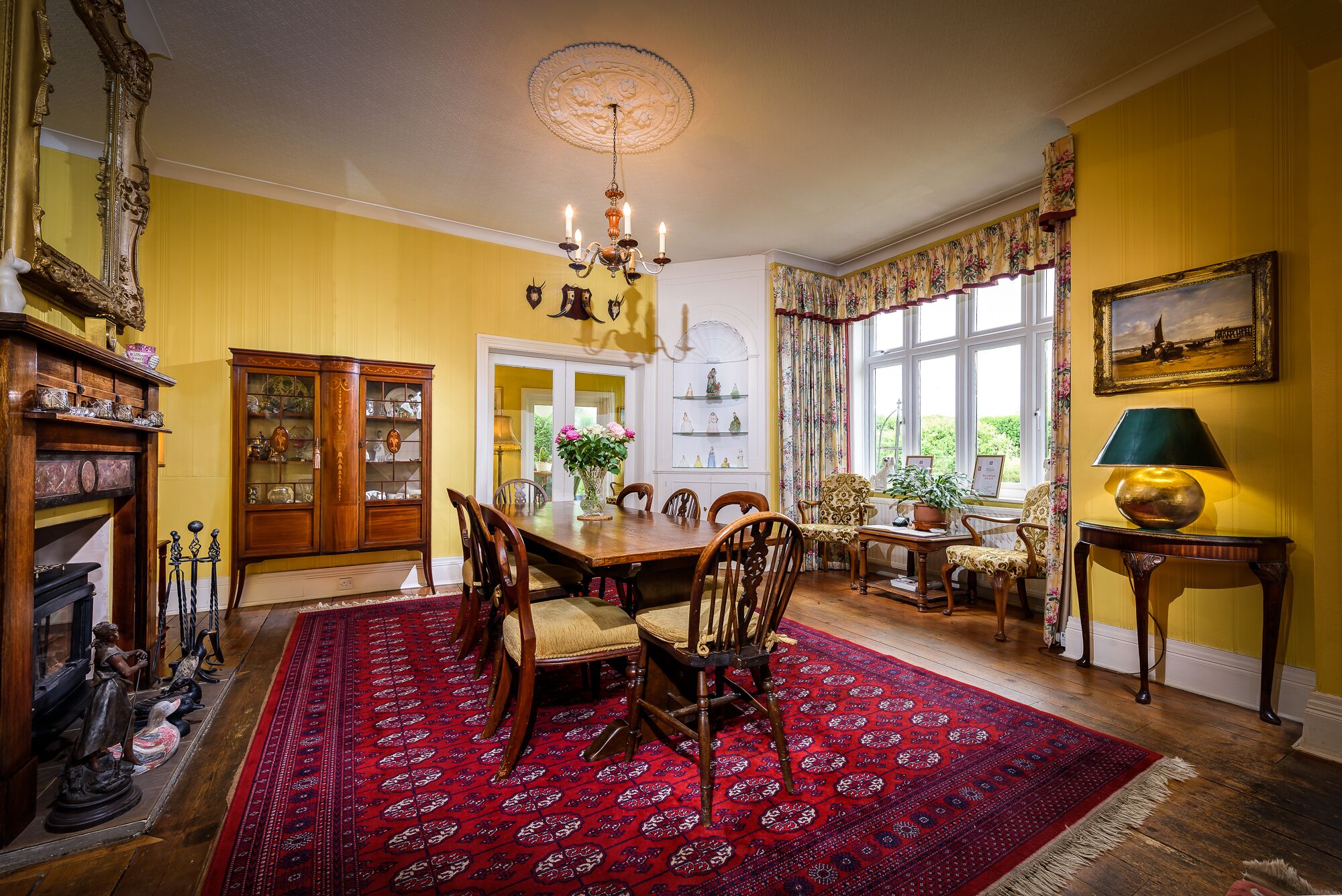 A dining room with a large wooden table, chairs, a red patterned rug, a chandelier, and a window with floral curtains.
