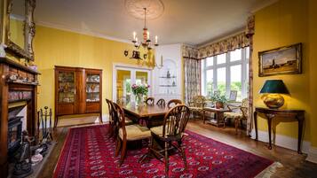 A dining room with a large wooden table, chairs, a red patterned rug, a chandelier, and a window with floral curtains.