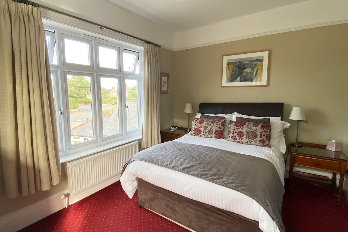 A hotel room with a large bed, a window with curtains, a painting on the wall, and a nightstand with a lamp.
