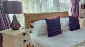 A neatly made bed with white linens and two purple pillows, a bedside lamp, and a decorative vase on a white nightstand. - Two Beaches (Paignton)