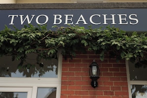Une enseigne portant l’inscription « TWO BEACHES » au-dessus d’un mur de briques agrémenté d’une fenêtre et d’une lanterne noire.